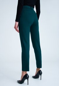 Zwieżone zielone spodnie wykonane z miękkiego materiału, z gładką fakturą, klasycznymi kieszeniami i wysokim stanem, w połączeniu z czarnymi szpilkami.