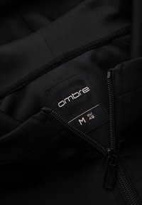 Veste noire avec fermeture éclair partiellement ouverte montrant l'étiquette de marque « ombre » et l'étiquette de taille marquée M, eur 48 à l'intérieur du col.