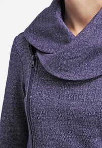 Sudadera gris y morada con cuello tipo cowl y cierre de cremallera. La tela tiene una apariencia texturizada y jaspeada. Vista en primer plano del escote.