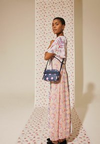 Ziedu maxi kleita ar īsām piedurknēm pasteļtoņos un tumši zila soma ar rozā punktiņiem. Modelis stāv priekšā ziedu fona.
