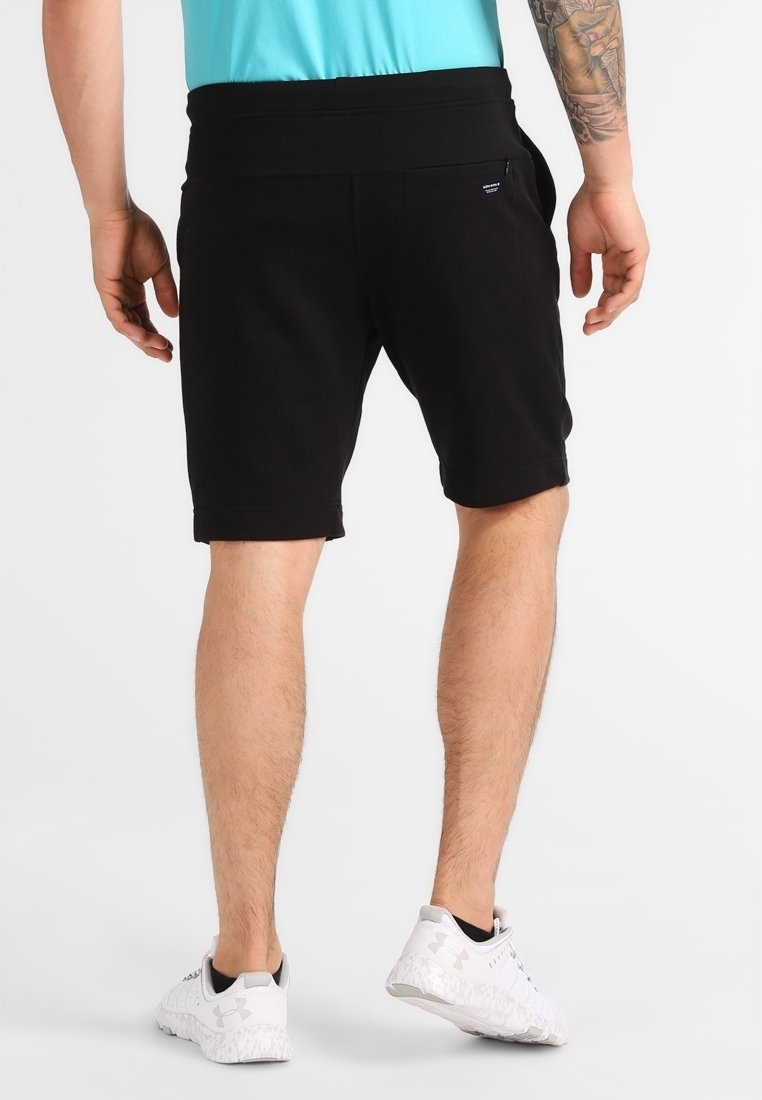 Shorts de sport noirs avec une large taille, des poches latérales et un ourlet roulé. Portés avec des chaussures de sport blanches.