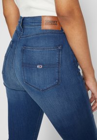 Vaqueros de mezquilla azul oscuro con una cintura alta, que presentan un parche de cuero en la parte posterior etiquetado "TOMMY JEANS" y dos bolsillos traseros con detalles de costura.