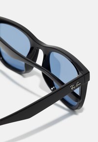 Ochelari de soare negri cu rame îndrăznețe, lentile de culoare albastră și marcaj Ray-Ban pe lateral. Materialul este lucios și din plastic.