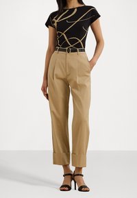 Czarny top z złotym abstrakcyjnym wzorem, zestawiony z tanimi spodniami o wysokiej talii z mankietami i czarnym paskiem, uzupełniony czarnymi sandałami na obcasie.