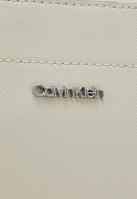 Biały teksturowany materiał z przeszyciami, z srebrnym logo "Calvin Klein". Projekt jest elegancki i minimalistyczny.