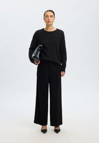 Suéter preto texturizado combinado com calças largas pretas. Modelo segura uma bolsa de couro preta com alça curvada. Roupa simples e monocromática.