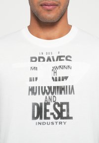 Bărbat îmbrăcat într-un tricou alb cu guler rotund, cu text tipărit negru și un logo alb parțial ascuns pe piept.