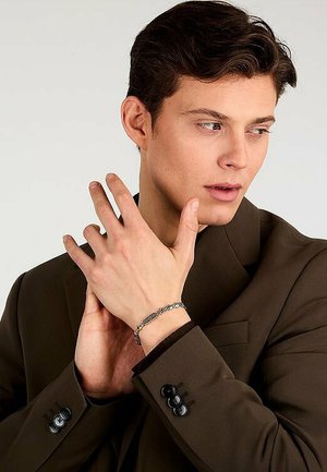 Strieborný náramok z retiazky s obdlžníkovými článkami a kruhovými akcentmi na zápästí modelky, ktorá má na sebe hnedý blazer s čiernymi gombíkmi.