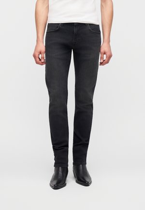 Άνδρας με στενά μαύρα τζιν, μαύρα μποτάκια με μυτερή μύτη και λευκό πουκάμισο, στέκεται μπροστά σε ουδέτερο φόντο.