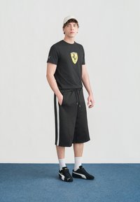 Νέος άνδρας που φοράει μαύρο T-shirt Ferrari, μαύρο σορτς με λευκές ρίγες, λευκό καπέλο, λευκές κάλτσες και μαύρα αθλητικά παπούτσια Puma, στέκεται σε μπλε χαλί.