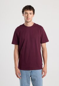 Young man with short curly hair wearing a plain burgundy t-shirt and light blue jeans, standing against a plain white background.