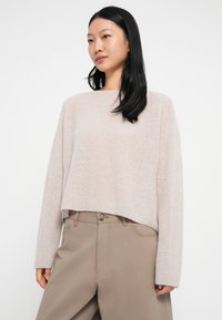 Femeie cu păr lung și negru, îmbrăcată cu un pulover moale de tricotaj bej deschis și pantaloni înalți de talie în nuanță de taupe, pe un fundal alb simplu.