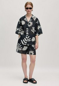 Schwarzes kurzärmliges Hemd und Shorts-Set mit großen weißen botanischen Mustern, aus leichtem Stoff, kombiniert mit schwarzen Slipper-Sandalen.