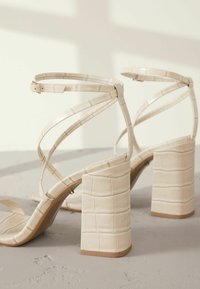 Beige High Heels Sandalen mit einem geprägten Krokodil-Muster, versehen mit schlanken Riemen und einem klobigen, rechteckigen Absatz. Glatte Oberfläche und verstellbare Schnalle.