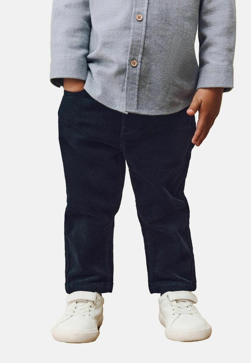 Enfant portant une chemise grise à manches longues boutonnée, un pantalon en velours côtelé noir et des baskets blanches à scratch, debout avec une main dans la poche.