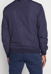 Granatowa kurtka bomberka ma gładką teksturę, prążkowane mankiety i dolną część oraz prosty design z centralnym szwem z tyłu.