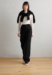 Pantaloni neri a vita alta e una maglietta aderente bianca a maniche lunghe, con un maglione nero appoggiato sulle spalle, abbinati a sneakers nere.