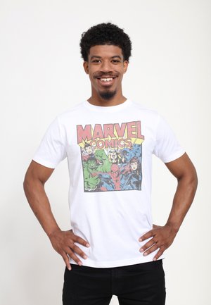 Homme souriant aux cheveux bouclés portant un t-shirt blanc de Marvel Comics représentant Hulk, Thor, Spider-Man et Black Widow, debout les mains sur les hanches.