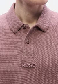 Μπλουζάκι πόλο σε ανοιχτό μωβ χρώμα, με υφή υφάσματος. Διαθέτει κλασικό γιακά, δύο κουμπιά και μεταλλικό λογότυπο "HUGO" στην αριστερή πλευρά.