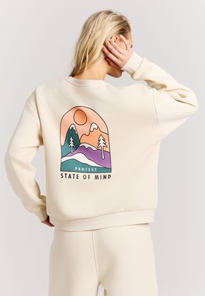 Blonde vrouw in crèmekleurige outfit met hand op het hoofd, draagt een sweatshirt met een kleurrijk bergdesign en de tekst "Protest State of Mind" op de achterkant.