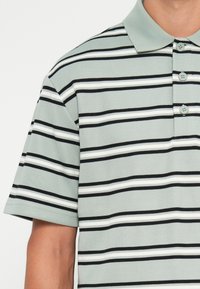 Korteermet polo-shirt laget av teksturert stoff, med horisontale striper i mintgrønn, hvit og svart, og tre knapper ved kragen.