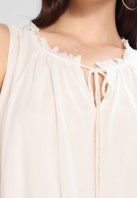 Nahaufnahme einer Person, die eine ärmellose weiße Bluse mit einem fransigen Ausschnitt und einer gebundenen Seilraffung an der Brust trägt.