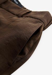 Brauner Stoff mit glatter Textur, ausgestattet mit einer Fronttasche und einem umgeschlagenen Bündchen-Design. Klassischer Stil mit minimalen Nähdetails.