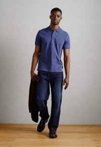 Polo shirt blu navy e jeans scuri. Il modello tiene un maglione bordeaux. Scarpe marroni. Design semplice, maniche corte e tessuto leggermente texture.