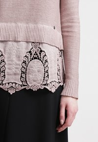 Maglione rosa chiaro con un design a maglia testurizzata e un orlo inferiore ricamato in pizzo. Le maniche a costine e il punto vita accentuano la silhouette.