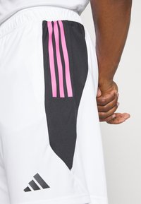 Gros plan d'un short de sport blanc avec des bandes noires et roses et une main reposant à côté de la cuisse.