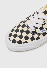 Czarno-biały w kratkę sneaker Vans z białymi sznurowadłami i białą gumową podeszwą, pokazujący górną przednią część i metkę marki.