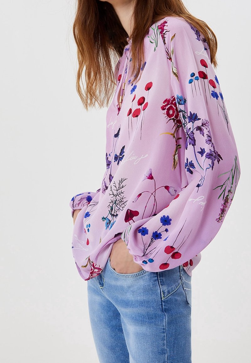 Ljuslila blus i mjukt tyg, med ett blommönster i röd, blå och grön, med långa ärmar och avslappnad passform.