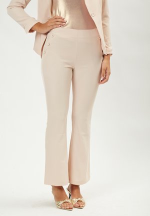 Light pink flared trousers with a smooth, matte texture and decorative buttons on the waistband, paired with metallic heeled sandals.