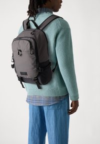 Sac à dos synthétique gris avec des accents noirs, bretelles réglables, poche avant et crochets, porté sur un pull en laine léger bleu clair et un jean ample.