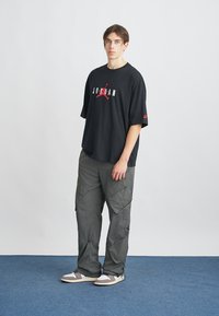 Črn bombažni T-shirt z grafiko "JORDAN" v beli in rdeči barvi. Ohlapne sivo cargo hlače z zadrgo in prilagodljivimi manšetami. Svetlo sivi superge.
