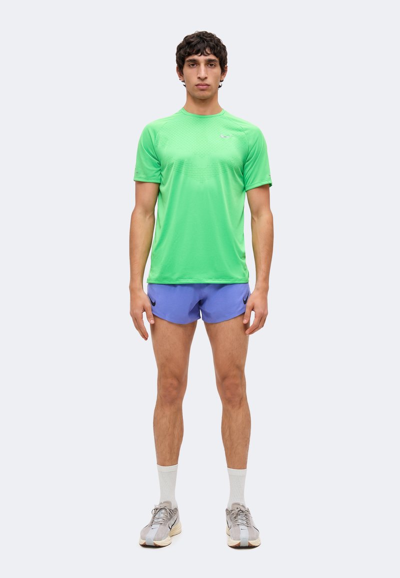 Man standing facing forward, wearing a bright green T-shirt, blue running shorts, white socks, and gray athletic shoes, on a plain background.