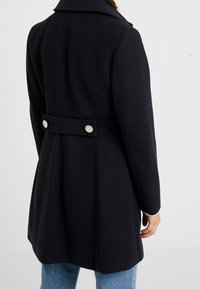 Cappotto di lana blu navy con ampio colletto, dettaglio della cintura con bottoni sulla schiena e texture liscia. Presenta due bottoni metallici in vita.