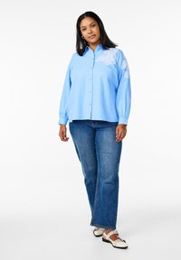 Camicia azzurra chiaro con maniche lunghe e ricamo floreale bianco sulla spalla sinistra, abbinata a jeans blu classici e scarpe nere.