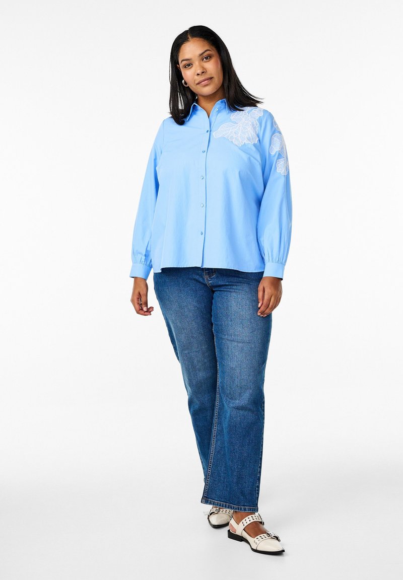 Camicia azzurra chiaro con maniche lunghe e ricamo floreale bianco sulla spalla sinistra, abbinata a jeans blu classici e scarpe nere.