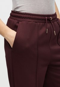 Μπορντό φόρμα με υφή ύφασμα, ελαστική μέση, κορδόνι με χρυσές λεπτομέρειες και πλαϊνές τσέπες.