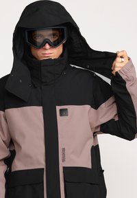 Zwarte en mauve waterdichte jas met een hoge kraag, grote voorzakken en logo-detail; gedragen met beschermende goggles.