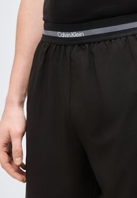 Κοντινό πλάνο ενός ατόμου που φοράει μαύρο σορτς με ελαστική μέση Calvin Klein και μαύρο πουκάμισο, με το χέρι χαλαρό στο πλάι.