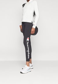 Svarta leggings med vitt "Champion"-logotyp, kombinerat med en långärmad vit polo. Skorna är vita och grå i atletisk stil.