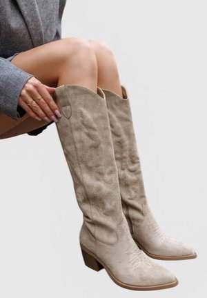 Personne portant des bottes de cow-boy en daim beige montant jusqu'aux genoux, avec des talons carrés, assise les jambes croisées et la main posée sur la botte gauche.