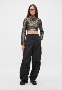 Μεταλλικό κομμένο μπουφάν με λεπτομέρειες από κουμπιά, σε συνδυασμό με φαρδιά μαύρα παντελόνια cargo με τσέπες και υφή. Μαύρα πλατφόρμα παπούτσια.