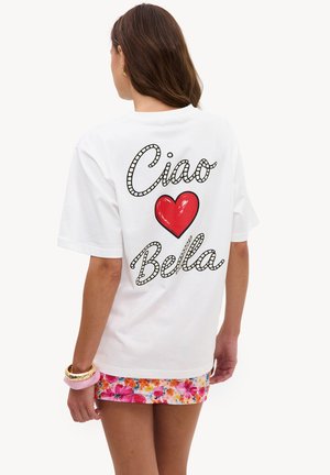 Femme portant un t-shirt blanc avec "Ciao Bella" et un cœur rouge dans le dos, associé à une jupe fleurie colorée et des bracelets.