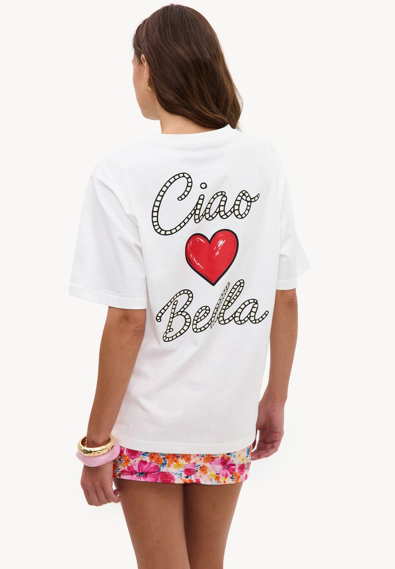Femme portant un t-shirt blanc avec "Ciao Bella" et un cœur rouge dans le dos, associé à une jupe fleurie colorée et des bracelets.