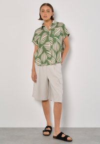 Camicia a maniche corte con colletto a pattern verde a foglie, abbinata a pantaloni corti beige e sandali neri. Tessuto morbido e vestibilità rilassata.