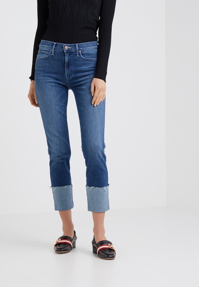 Blue denim jeans with a fitted design, cropped length, and frayed light blue cuffs. Paired with black shoes featuring a red strap and studs.