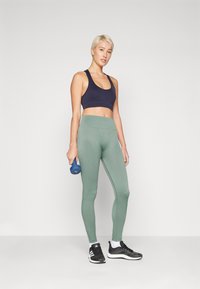Sports BH i marineblå, høytlivede grønne leggings med ribbet tekstur, og svarte joggesko. Holder en blå manual i høyre hånd.